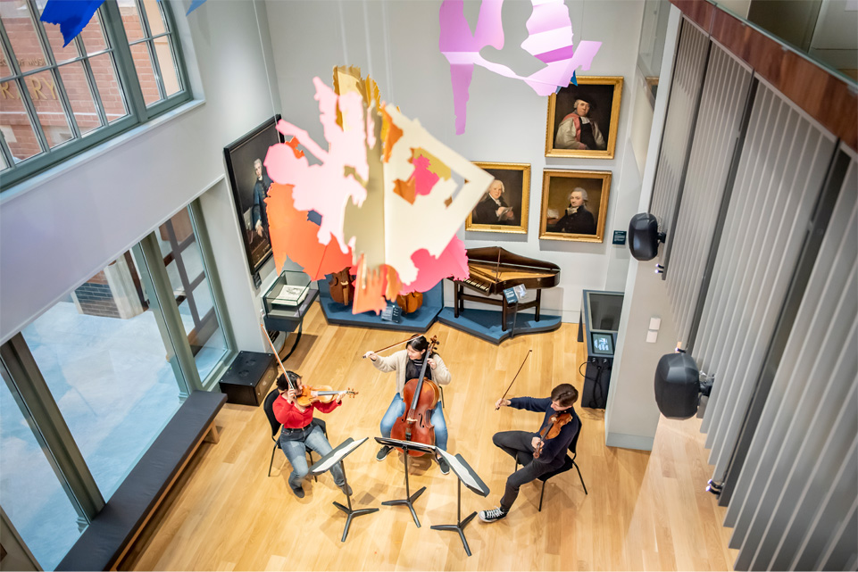 Two violinists and one cellist, one male and two female, perform music on the ground floor of the museum, seated under a sculptural artwork descending from the ceiling.